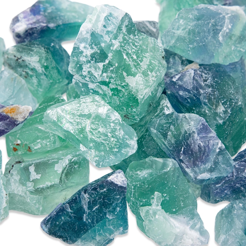 May include: A collection of raw fluorite crystals in shades of green, blue, and purple. The rough, natural stones have a translucent quality, with varying depths of color and visible internal structures. The crystals are piled together against a white background.