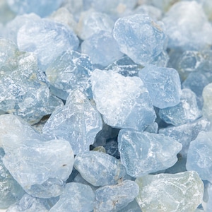 May include: A close-up shot of a pile of light blue rough-cut gemstones. The stones vary in size and shape, with a crystalline appearance. The color is a soft, pale blue, and the overall effect is cool and calming.