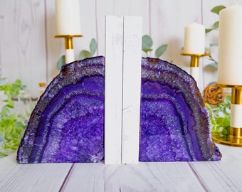 May include: A pair of bookends featuring two halves of a deep purple agate geode slice. Each piece has a rough, crystalline interior and a smooth, polished outer edge, attached to a vertical white wooden support for stability on a flat surface.