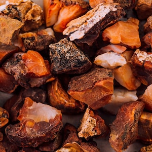 May include: A close-up of a pile of rough, orange, red, and brown carnelian stones. The stones are various sizes and shapes.