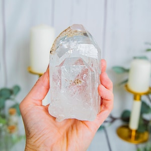 May include: A clear, translucent quartz crystal point with a hexagonal structure and natural internal inclusions. The crystal features a pointed termination and a rough, uneven base, displaying varying degrees of transparency and light refraction.