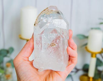 May include: A clear, translucent quartz crystal point with a hexagonal structure and natural internal inclusions. The crystal features a pointed termination and a rough, uneven base, displaying varying degrees of transparency and light refraction.
