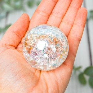 May include: A clear, iridescent crystal ball with a cracked, textured surface is held in an open hand. The sphere reflects light, displaying a range of colors. The background is blurred, with hints of greenery and a light-colored wooden surface.