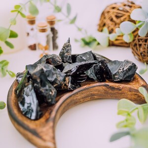 Puede incluir: Una colecci&oacute;n de cristales de obsidiana negra en una bandeja de madera en forma de media luna. Los cristales son crudos y tienen una textura natural y rugosa.