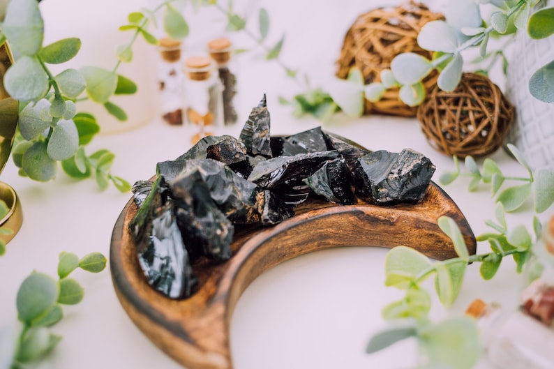 Puede incluir: Una colecci&oacute;n de cristales de obsidiana negra en una bandeja de madera en forma de media luna. Los cristales son crudos y tienen una textura natural y rugosa.
