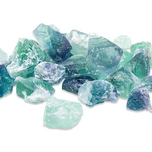 May include: A collection of raw fluorite crystals in shades of green, blue, and purple. The crystals have a rough, natural texture and varying shapes, creating a visually appealing display. The gemstones are scattered against a white background.