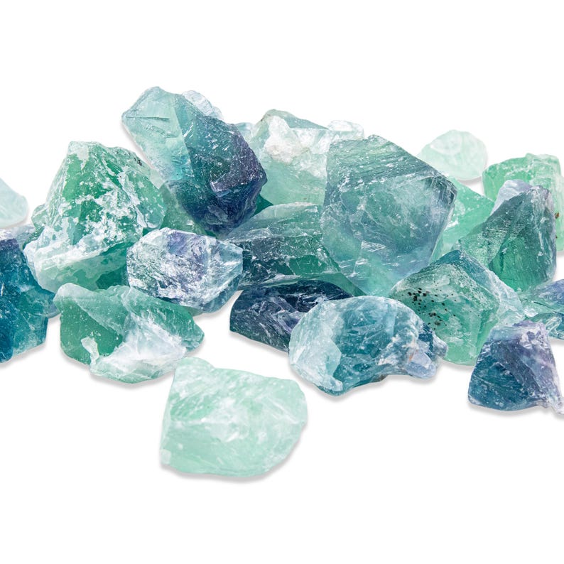 May include: A collection of raw fluorite crystals in shades of green, blue, and purple. The crystals have a rough, natural texture and varying shapes, creating a visually appealing display. The gemstones are scattered against a white background.