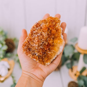May include: A large, rough, orange and yellow citrine crystal cluster in a hand. The crystal has a natural, raw, and unpolished look.