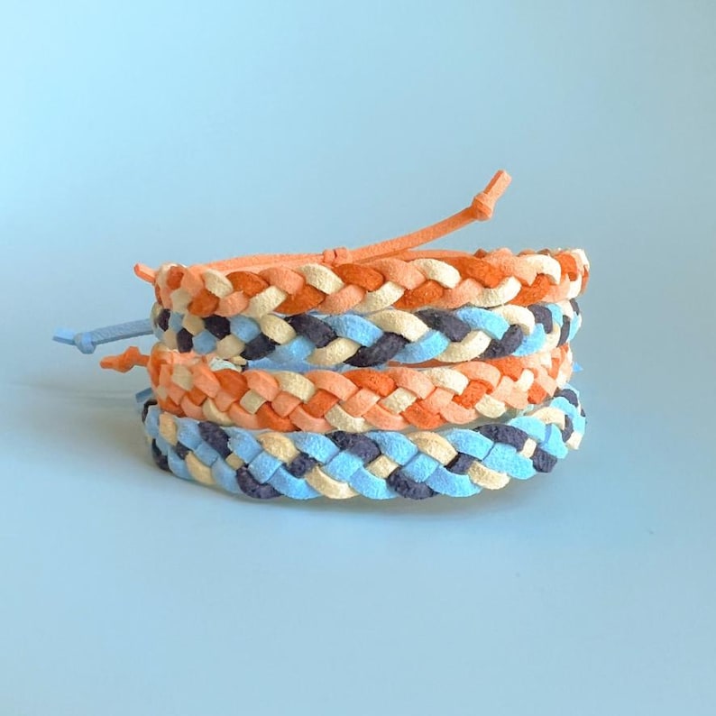 May include: A stack of four braided bracelets in shades of orange, cream, blue, and navy. Each bracelet has a matching adjustable tie closure. The bracelets are stacked on a light blue background.