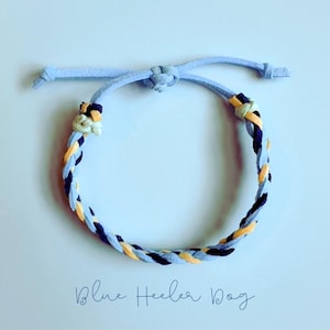 May include: A blue braided bracelet with yellow and navy accents. The bracelet has a light blue adjustable cord. The text "Blue Heeler Dog" is written in cursive at the bottom of the image.