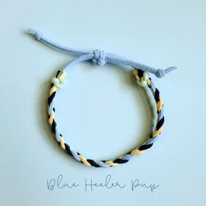 May include: A braided bracelet with a light blue suede cord and a woven band of black, yellow, and light blue. The bracelet is adjustable with a sliding knot closure. The text "Blue Heeler Pup" is written in cursive at the bottom.