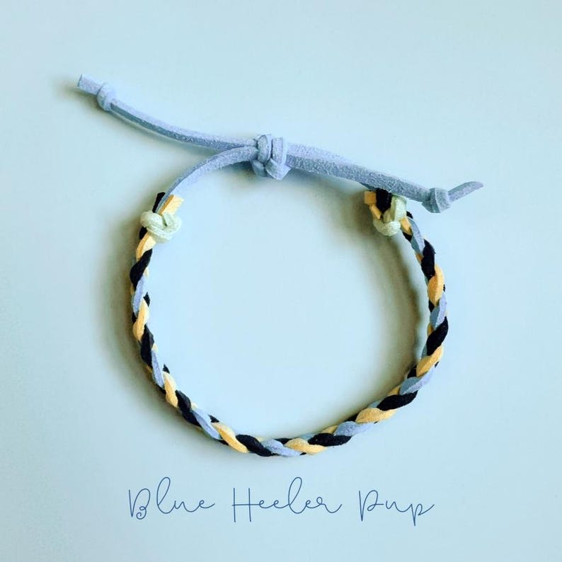 May include: A braided bracelet with a light blue suede cord and a woven band of black, yellow, and light blue. The bracelet is adjustable with a sliding knot closure. The text "Blue Heeler Pup" is written in cursive at the bottom.