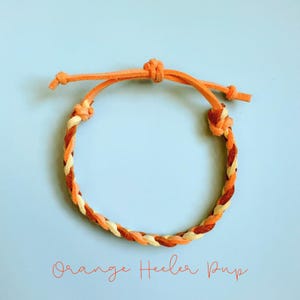 May include: Orange and cream-colored braided bracelet with adjustable ties. The bracelet features a woven design with orange, cream, and brown strands. The text "Orange Heeler Pup" is written in orange script below the bracelet.