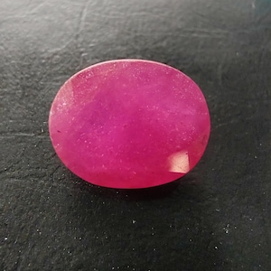 May include: A vibrant, oval-shaped, hot pink gemstone with a faceted surface. The stone displays a translucent quality with internal inclusions and a polished finish, reflecting light across its rounded edges and flat top surface.