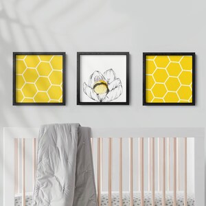 Può includere: Tre stampe incorniciate con un motivo a nido d'ape giallo, un'illustrazione in bianco e nero di un fiore con un'ape e un altro motivo a nido d'ape giallo. Le stampe sono appese a una parete bianca sopra una culla bianca con una coperta grigia.