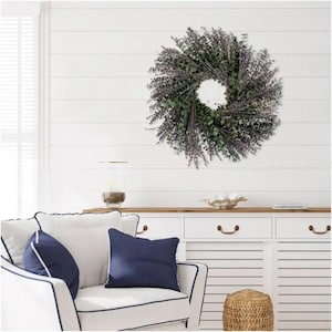 May include: A green and purple eucalyptus wreath hanging on a white shiplap wall. The wreath is in a living room setting with a white couch, blue pillows, a wooden cabinet, and a woven basket.