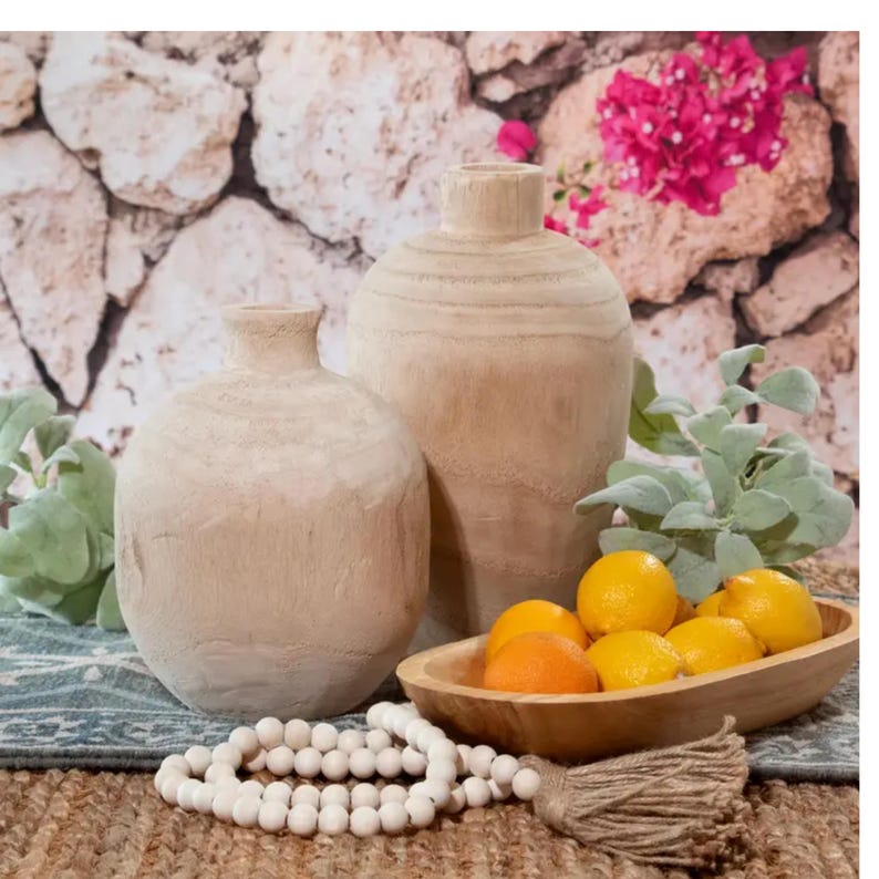May include: Two wooden vases, one tall and one short, with a light brown finish. The vases are sitting on a woven rug with a wooden bowl filled with lemons.