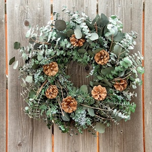 May include: A green eucalyptus wreath with brown pine cones. The wreath is made of fresh greenery and is decorated with pine cones.