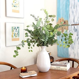 May include: A white ceramic vase with a rounded base and a narrow neck holds a bouquet of green eucalyptus leaves. The vase is sitting on a wooden table with a stack of books and a candle.