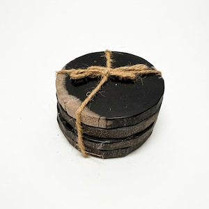 May include: A stack of four circular coasters featuring a smooth, glossy black top surface and a textured, natural dark brown edge. The coasters are bound together by a thin, light brown jute twine tied in a simple cross pattern at the top.