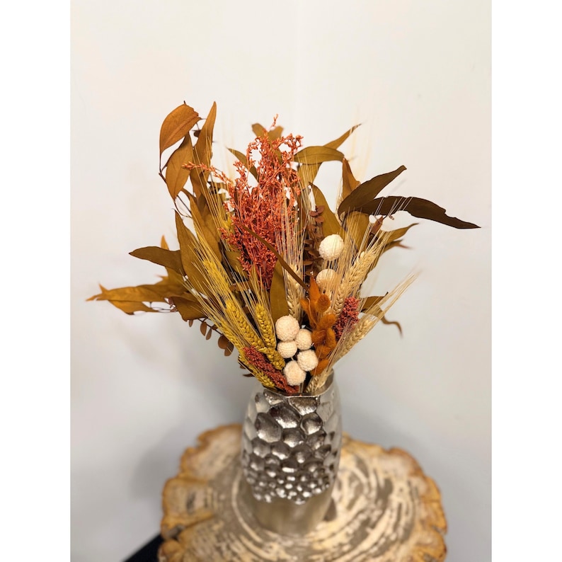 May include: A dried flower arrangement in a silver vase with a textured surface. The arrangement features dried grasses, wheat, and small white and orange flowers.