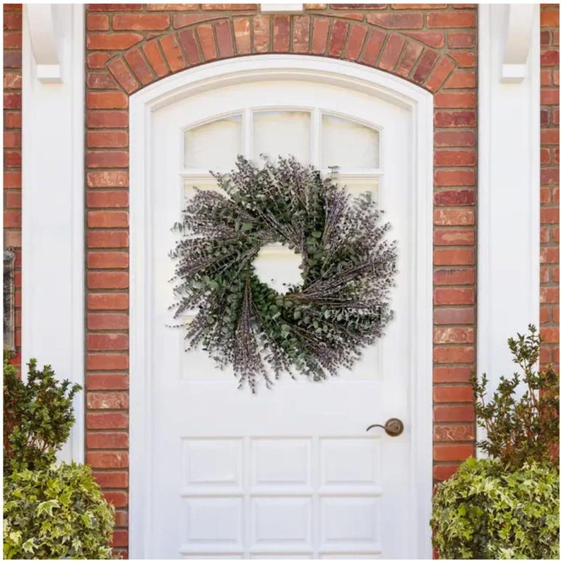 May include: A green and purple eucalyptus and lavender wreath hangs on a white front door with a white door frame. The door is surrounded by red brick.