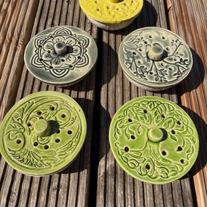 Könnte beinhalten: Fünf kreisförmige Keramikdeckel mit zentralen Knöpfen und verschiedenen geprägten Mustern. Ein Deckel ist leuchtend gelb, zwei sind dunkelgrau mit Blumen- und geometrischen Mustern, und zwei sind hellgrün mit Baum- und Vogelmotiven mit kleinen Löchern.