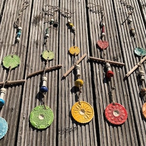 Könnte beinhalten: Handgefertigte Windspiele aus Keramik mit Blatt- und Kreisdesigns. Jedes Windspiel hat ein einzigartiges Farbschema und ist an Holzelementen und Schnur aufgehängt, was eine rustikale Ästhetik erzeugt. Maße in Zentimetern.