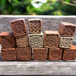 Könnte beinhalten: Eine Ansammlung kleiner, quadratischer Terrakotta-Blöcke mit verschiedenen geprägten Blumen- und geometrischen Mustern. Die Blöcke sind auf einer Holzoberfläche gestapelt und zeigen die detaillierten Muster und Erdtöne des Tons.