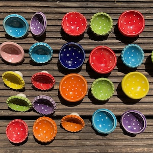 Könnte beinhalten: Eine Sammlung farbenfroher, pilzförmiger Keramikschalen mit weißen Tupfen. Die Schalen sind in verschiedenen Farben erhältlich, darunter Rot, Blau, Grün, Orange, Gelb und Lila. Die Schalen sind auf einer Holzoberfläche angeordnet.