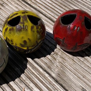 Könnte beinhalten: Vier Keramikkugeln mit Gesichtern im Jack-Skellington-Stil. Die Kugeln sind weiß, gelb und rot, mit genähten Mündern und ovalen Augenausschnitten. Sie liegen auf einer verwitterten Holzoberfläche.
