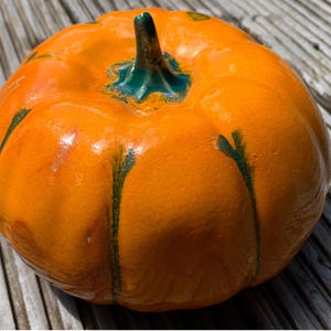 Könnte beinhalten: Ein glänzender, orangefarbener Keramik-Kürbis mit einem grünen Stiel und grünen vertikalen Streifen. Der Kürbis ist rund und hat eine gerippte Textur, ideal für Herbstdekorationen oder Halloween-Ausstellungen.