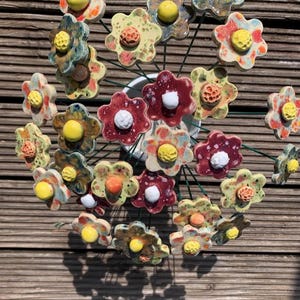 Könnte beinhalten: Eine handgefertigte Keramik-Blumenskulptur mit mehreren bunten Blumen. Die Blumen haben gelbe, weiße und rote Akzente mit grünen Stielen. Die Skulptur steht vor einem hölzernen Hintergrund.