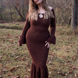 Puede incluir: Vestido largo de ganchillo marrón con mangas largas y escote de ojo de cerradura. El vestido tiene un corpiño ajustado y una falda acampanada. La modelo está de pie en una zona boscosa con hojas caídas en el suelo.
