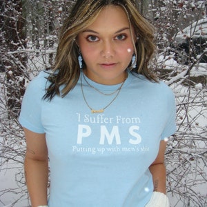 Könnte beinhalten: Hellblaues T-Shirt mit dem Text "I Suffer From PMS Putting up with men's shit" in Weiß. Die Person trägt eine goldene Halskette und weiße Fingerhandschuhe. Der Hintergrund ist ein verschneiter Wald.