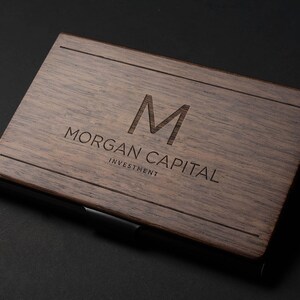 May include: A wooden business card holder with a dark brown finish. The holder has a metal clip and the text "MORGAN CAPITAL INVESTMENT" is engraved on the front.