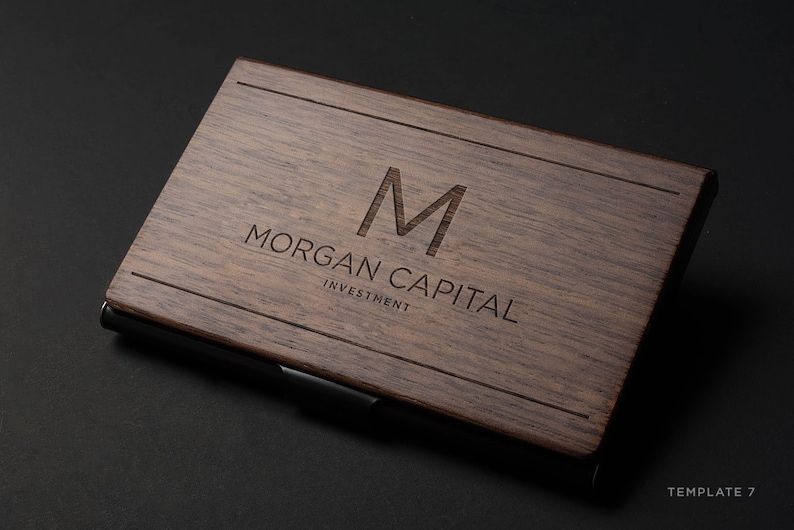 May include: A wooden business card holder with a dark brown finish. The holder has a metal clip and the text "MORGAN CAPITAL INVESTMENT" is engraved on the front.