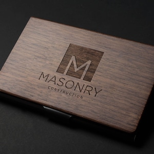 May include: A wooden business card holder with a black metal clip. The holder has a laser-engraved logo with the letter "M" inside a square and the text "MASONRY CONSTRUCTION".