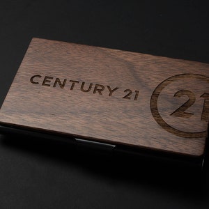May include: A wooden business card holder with a black metal trim. The holder has the text "CENTURY 21" and a circle with the number "21" engraved on the front.