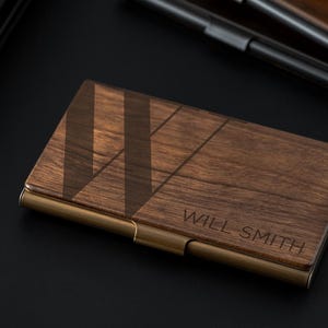 May include: A wooden business card holder with a dark brown finish and a geometric design. The holder has a brass-colored metal frame and the name "WILL SMITH" is engraved on the front. The product is on a black surface.