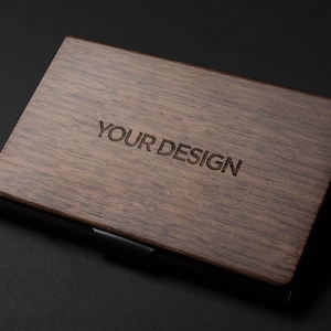 May include: A dark brown wooden business card holder with a black metal trim. The words "YOUR DESIGN" are engraved on the front.