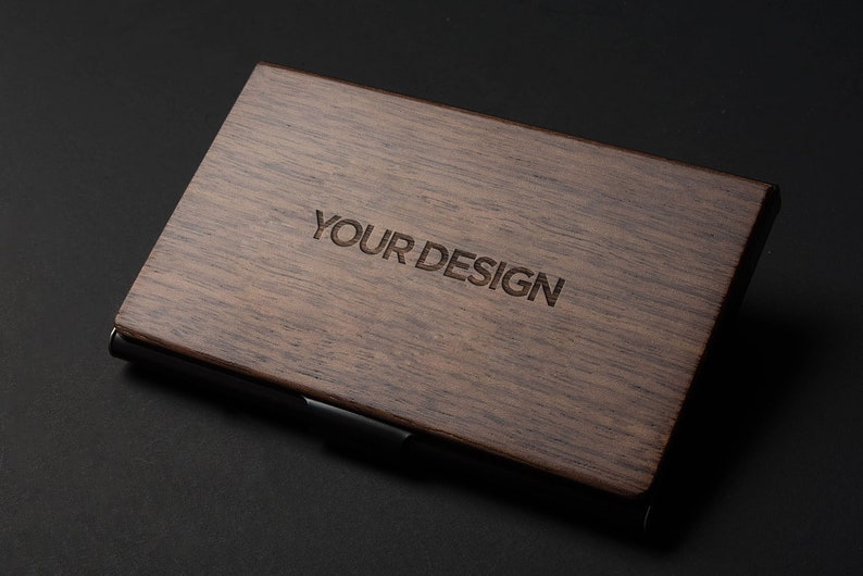 May include: A dark brown wooden business card holder with a black metal trim. The words "YOUR DESIGN" are engraved on the front.