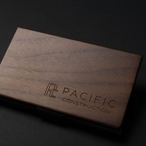 May include: A wooden business card holder with a metal edge. The holder has the logo "PC PACIFIC CONSTRUCTION" engraved on the front.