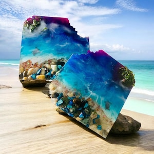 May include: Two square resin art pieces with a beach scene. The resin is clear and blue with white swirls and embedded seashells, stones, and sand. The pieces are on a wooden surface.