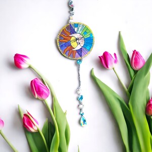 May include: A sun and moon stained glass ornament with a crystal bead chain. The ornament is hanging from a white ceiling and surrounded by pink tulips.