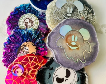 Puede incluir: Una colección de posavasos de resina hechos a mano con varios diseños. Los posavasos presentan imágenes de Mickey Mouse, Jack Skellington y la palabra "Boo". Los colores incluyen azul, morado, rosa y dorado. Los posavasos tienen bordes irregulares.