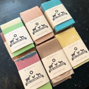 May include: A collection of rectangular goat milk soap bars in various colors including green, pink, blue, yellow, and brown. Each bar is wrapped in a tan paper band labeled with The Tucker Farm logo, a goat, chicken, and dog icon, and Net weight: 5 oz.