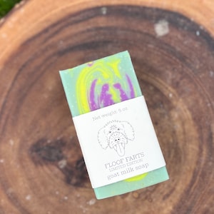 May include: A bar of goat milk soap with a green base and swirled yellow and purple colours. The soap is wrapped with a white label that reads "FLOOF FARTS LIMITED EDITION goat milk soap" and the net weight is 142 grams.