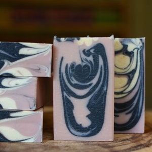 May include: Artisan soap bars with a pink base and swirling black and white patterns. The rectangular bars are stacked, displaying the marbled designs. The soap is likely scented and suitable for personal care, presented on a wooden surface.