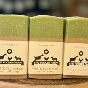 May include: Three rectangular bars of goat milk soap with a two-tone design featuring a light green top layer and a tan bottom layer. Each bar has a paper label printed with "The Tucker Farm," "White Tea & Pear," and "Net weight: 5 oz."
